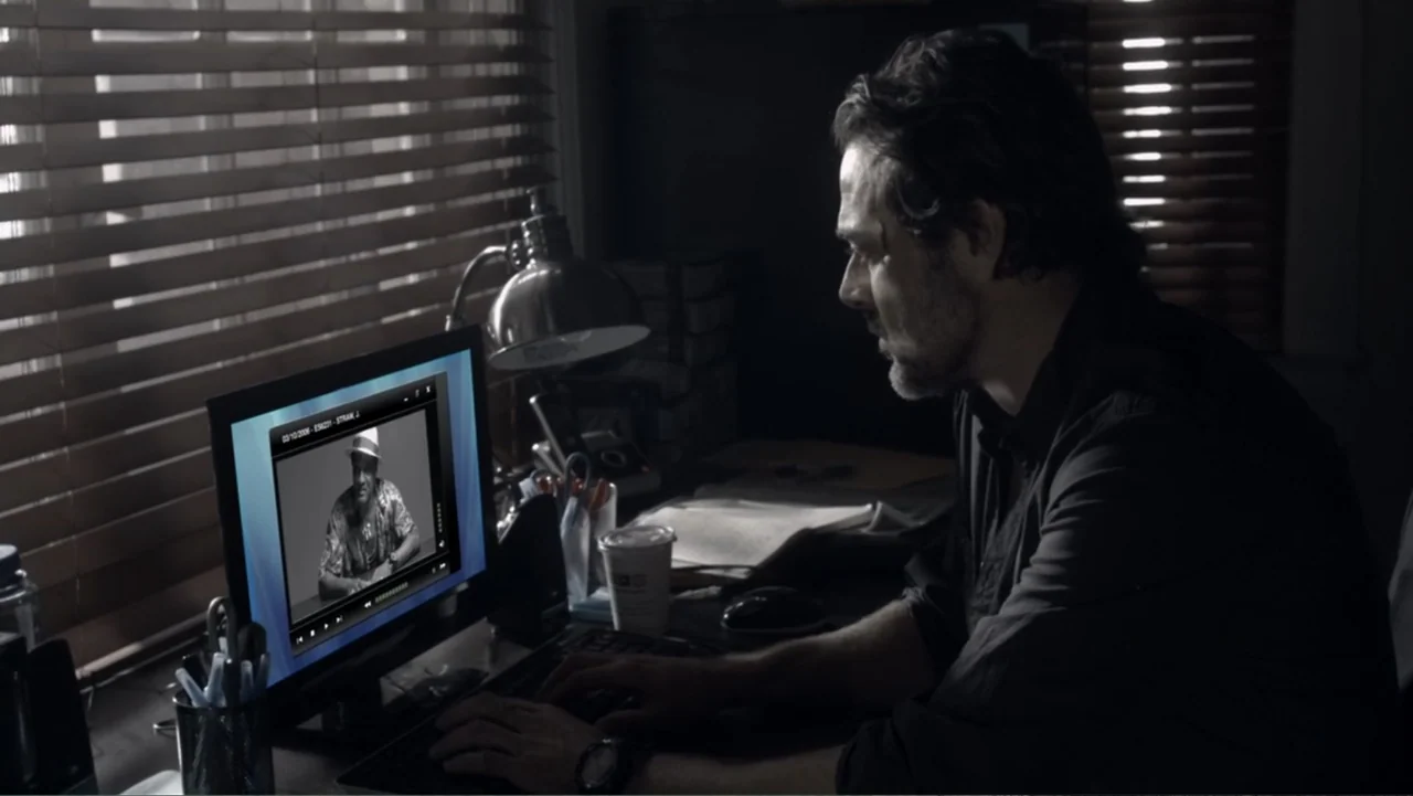 The Messenger (2015) movie still: medium — A dimly lit home office with horizontal window blinds and a cluttered desk. A man works at his computer…
