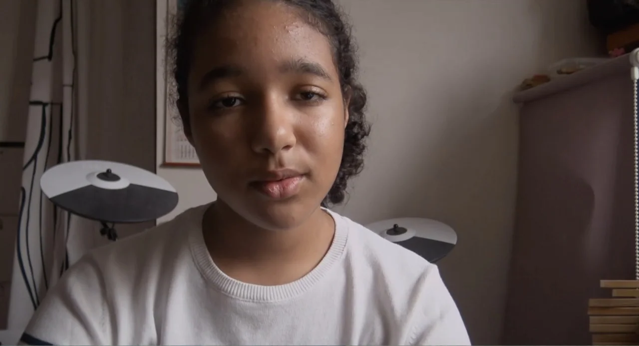 A Dramatic Film (2019) movie still: closeup — A teenage girl sits in a bedroom with electronic drum pads visible behind her. She faces the camera…