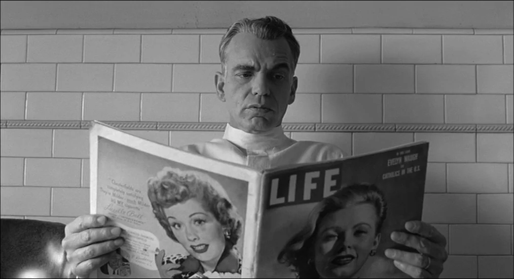 The Man Who Wasn’t There (2001) movie still: medium — A tiled wall serves as the background for a man holding a magazine. The man sits in front of…