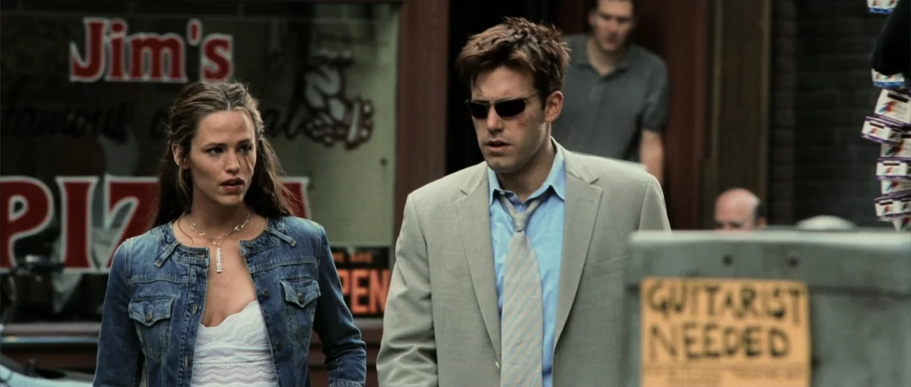 Daredevil (2015) movie still: medium — An urban sidewalk outside a pizzeria with a prominent window sign in the background. A woman and a man in a…