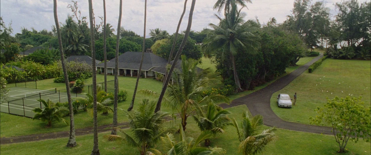 The Descendants (2011) movie still: establishing — A sprawling tropical property features a single-story house with a dark roof and lawn. A tennis…