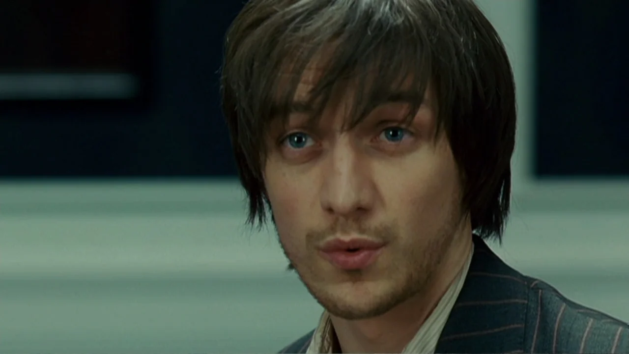 Penelope (2007) movie still: closeup — A man stands in an office environment with indistinct dark panels in the background. He is speaking, looking…