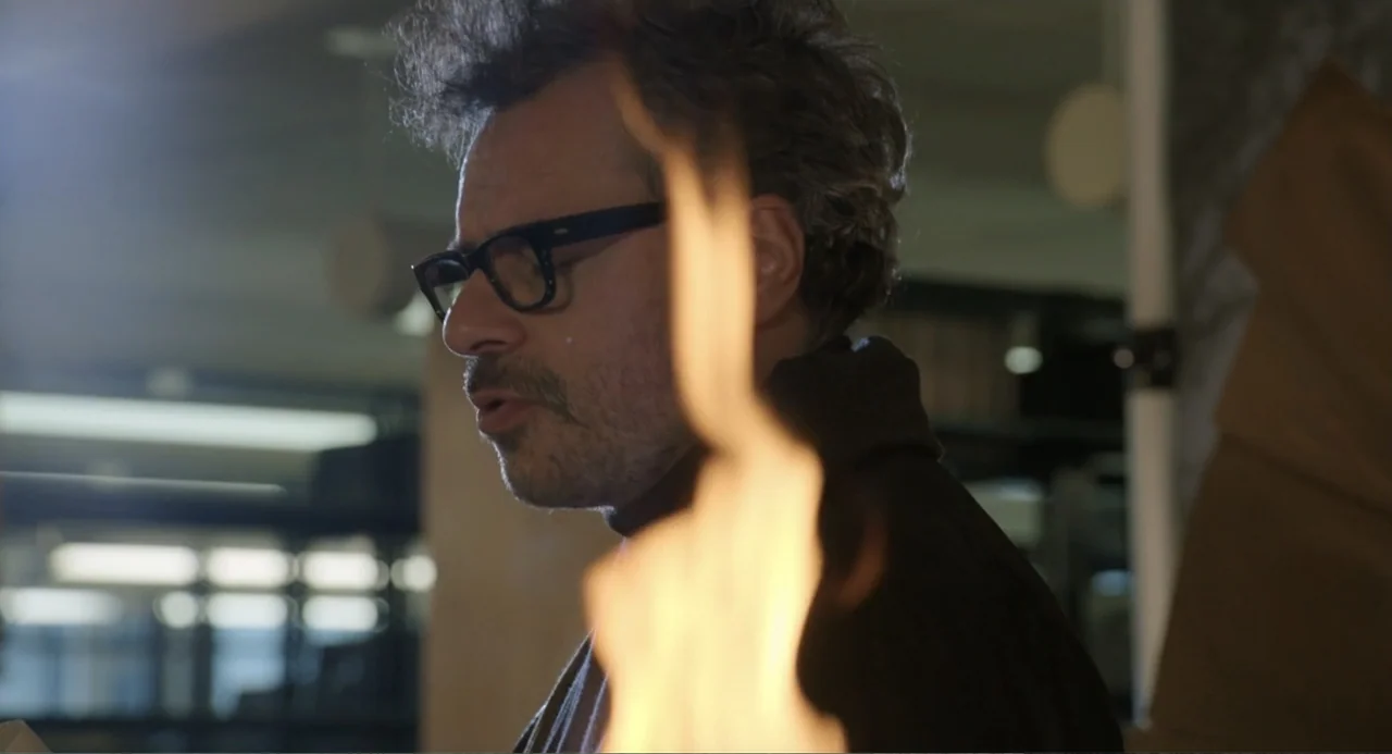 Harold and the Purple Crayon (2024) movie still: closeup — A dark indoor setting features a man in focus near a blurred flame. The subject stands…