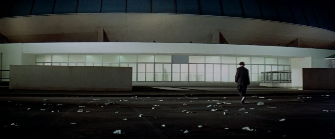 Soylent Green (1973) movie still: wide — A vast, dark paved lot sits before a stark, mid-century concrete stadium entrance. A lone man in a dark…