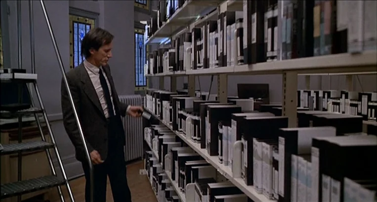 Videodrome (1983) movie still: medium — A quiet archive room features long rows of shelves filled with white and black binders. A man in a business…