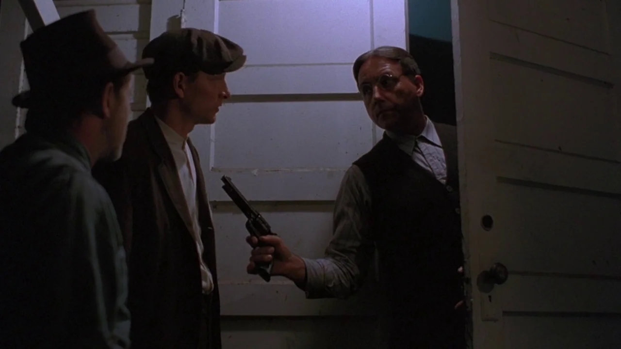 Matewan (1987) movie still: medium — A dark wooden doorway frames a man holding a revolver toward two visitors. The setting is dimly lit with white…