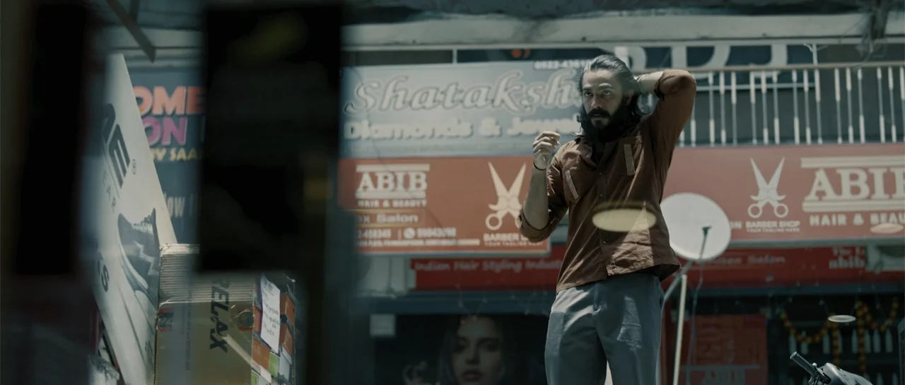 Nishaanchi 2 (2025) movie still: medium — An urban exterior features a man standing in front of commercial storefront signage. The man adjusts his…