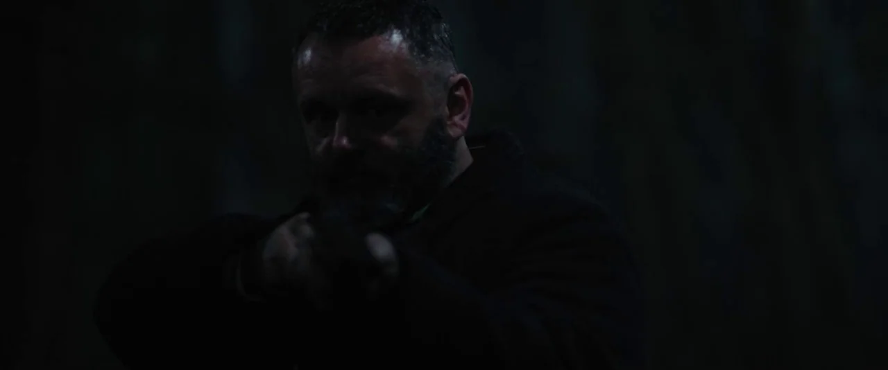 Apostle (2018) movie still: medium — A man stands in an undifferentiated dark outdoor space at night. He is pointing a firearm directly toward the…