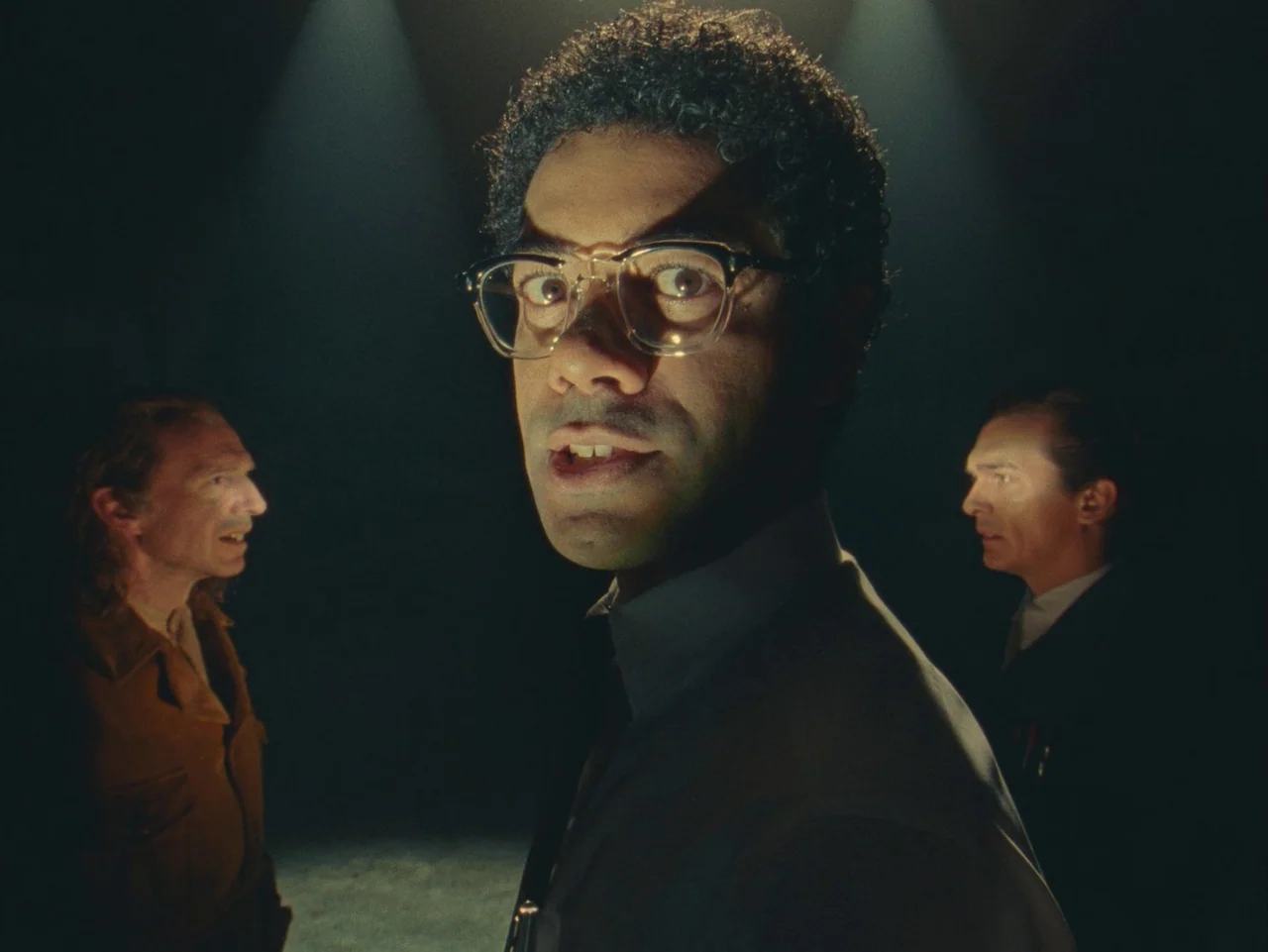 The Rat Catcher (2023) movie still: closeup — A dark, void-like space serves as a minimalist theatrical stage. A central man in glasses addresses…