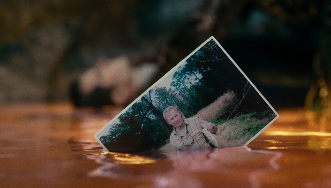 Justice League (2017) movie still: closeup — A glossy photograph shows a man fishing and sits in a shallow red liquid. The background is blurred and…