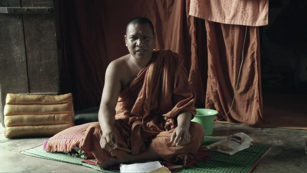 A Cambodian Spring (2018) movie still: medium — A sparsely furnished monastic interior with a concrete floor and draped cloth walls. A man in…