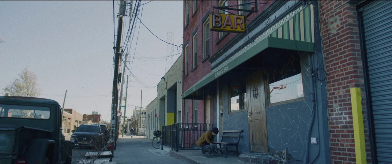 She Came To Me (2023) movie still: wide — A brick storefront marked as a bar sits on an urban neighborhood sidewalk. A person crouches beside a…