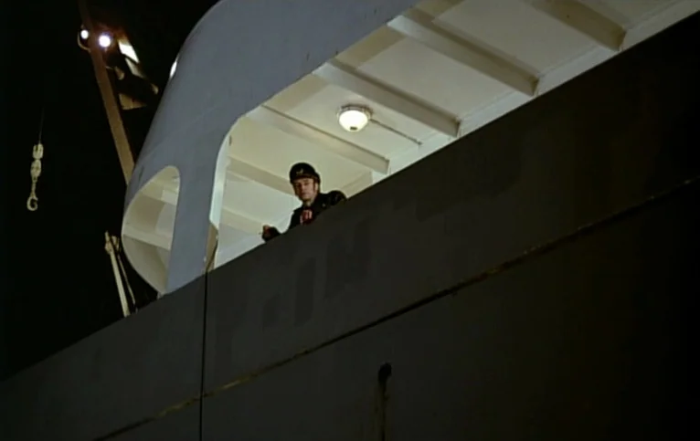 Giliap (1975) movie still: wide — The exterior of a ship's structure at night under an overhead artificial light. A man in a naval cap leans against…