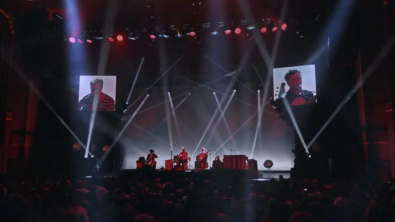 Flight of the Conchords: Live in London (2018) movie still: wide — A dimly lit concert hall stage features three musicians performing under multiple…