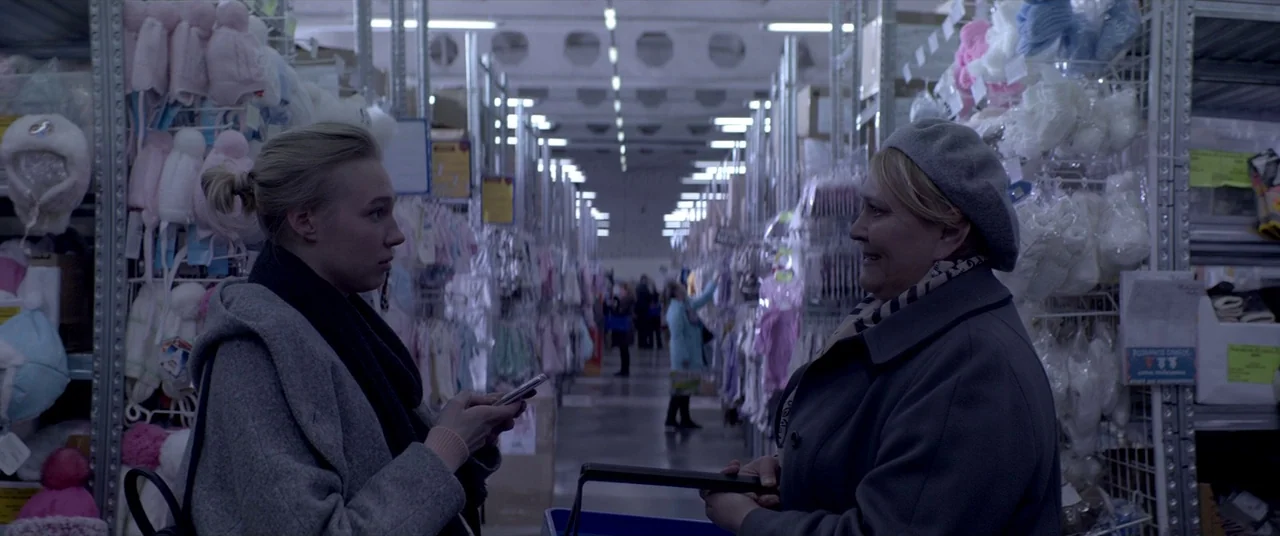 Loveless (2017) movie still: medium — A long retail aisle is lined with metal shelves packed with assorted baby hats. Two women stand in the…