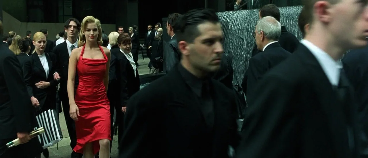 The Matrix (1999) movie still: fullbody — A busy urban plaza features a large decorative water wall in the background. A woman in a red dress walks…