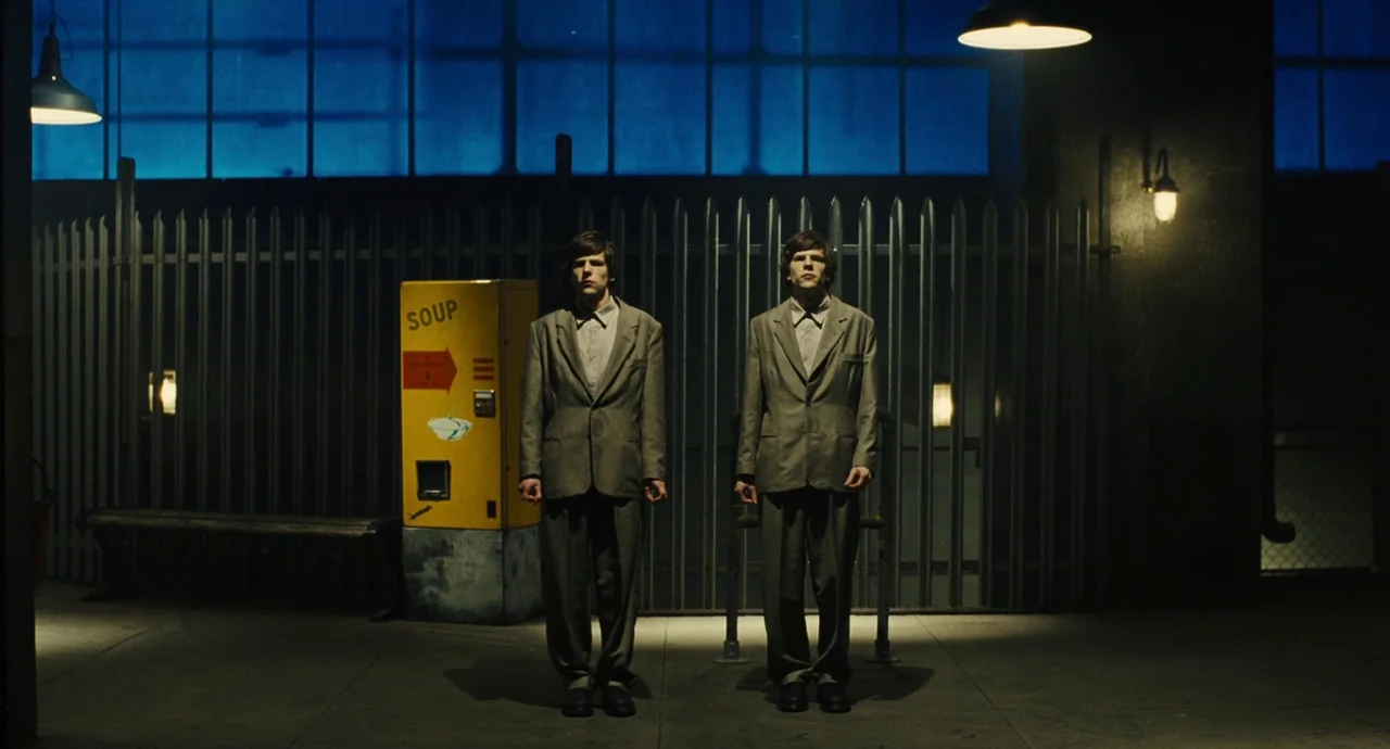The Double (2013) movie still: fullbody — An industrial space features a tall metal fence against a deep blue grid wall. Two identical men stand…