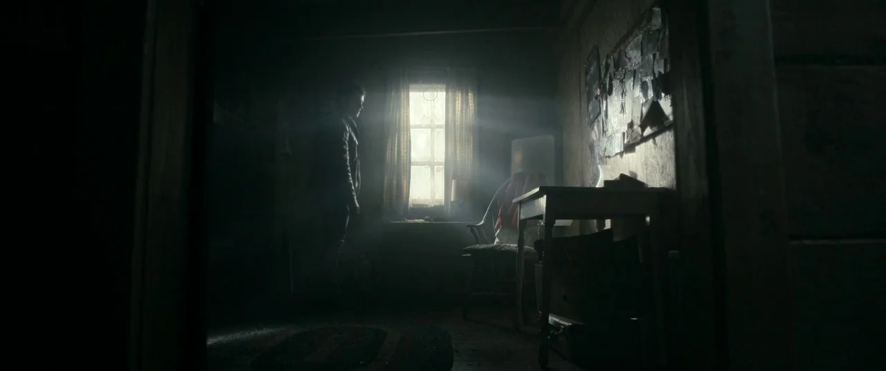 Evil Dead (2013) movie still: wide — A dimly lit room in a rustic cabin features wooden walls and scattered belongings. A man stands in silhouette…
