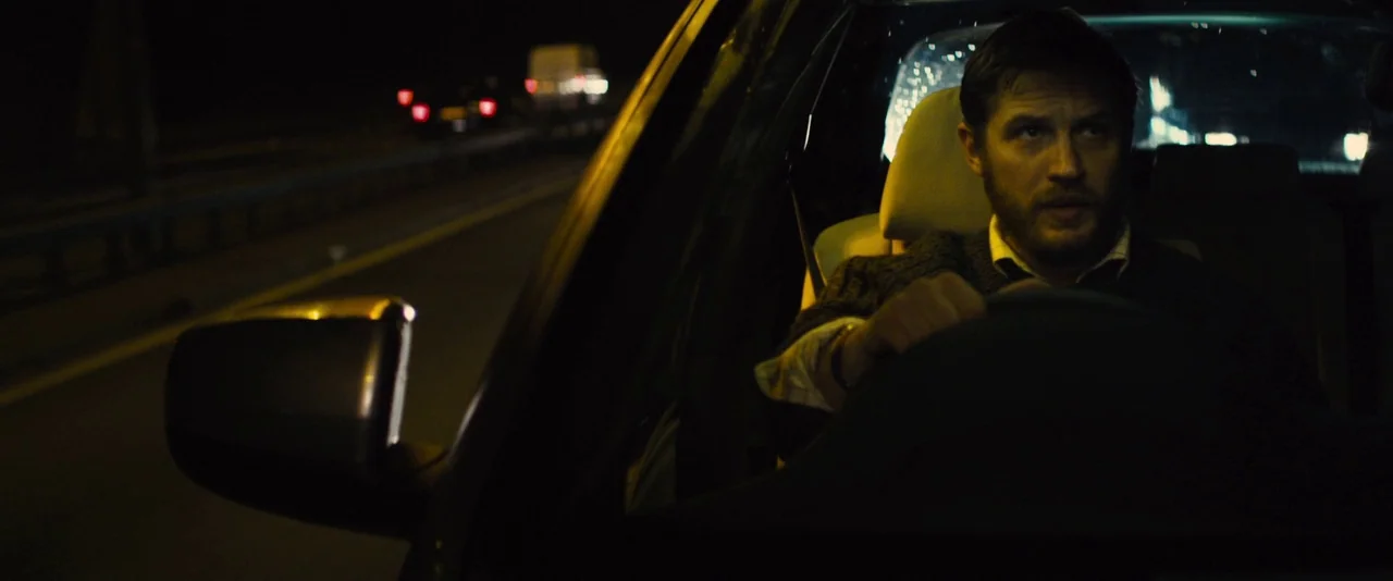 Locke (2013) movie still: medium — The interior of a moving car at night shows the driver behind the wheel. The man looks ahead while steering…