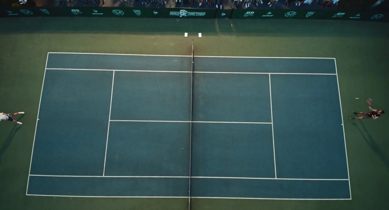 Challengers (2024) movie still: wide — A high-angle bird's-eye view of a professional tennis court at night. Two players stand at opposite sides of…