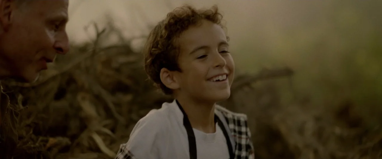 Winnie-the-Pooh: Blood and Honey 2 (2024) movie still: medium — A boy with curly hair laughs while standing outdoors in a rural setting. An adult…