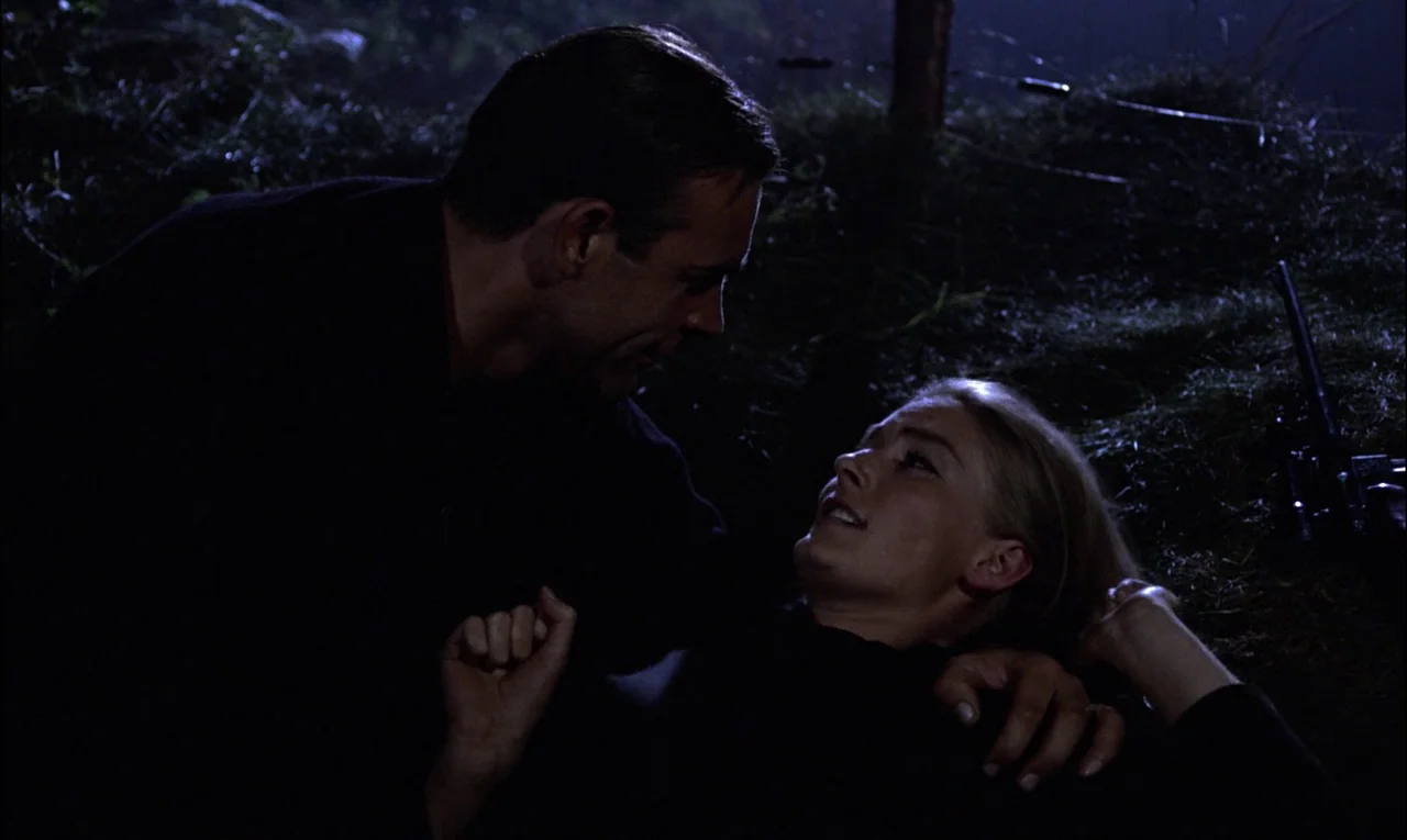 Goldfinger (1964) movie still: medium — A man and woman lie together in a grassy patch of dark ground. They are positioned close to each other in a…