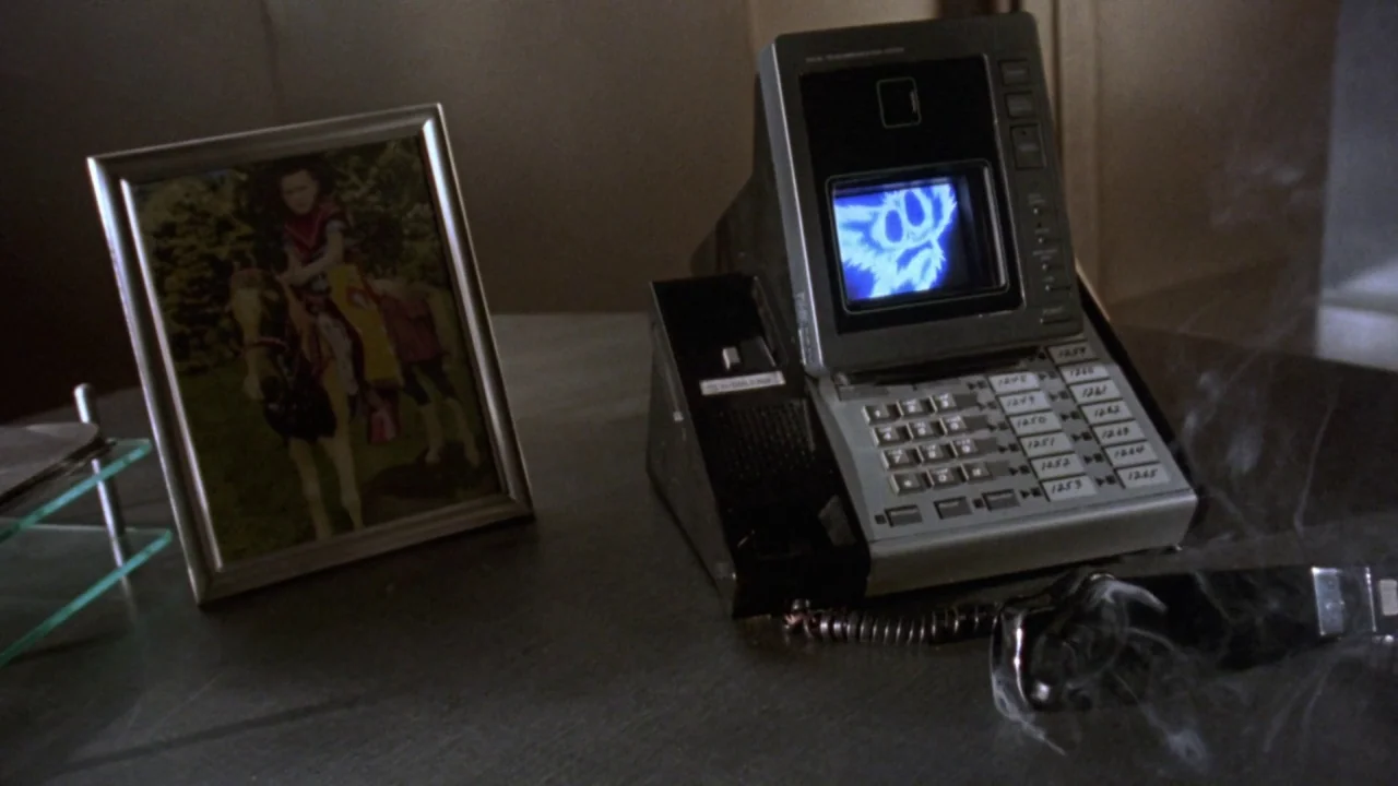 Gremlins 2: The New Batch movie still: closeup — A dark wooden desk surface holds a framed photograph and a retro digital telephone. Smoke rises…