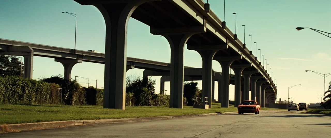 Green Lantern (2011) movie still: wide — A wide asphalt street stretches beneath a sprawling elevated highway overpass with concrete pillars. A red…