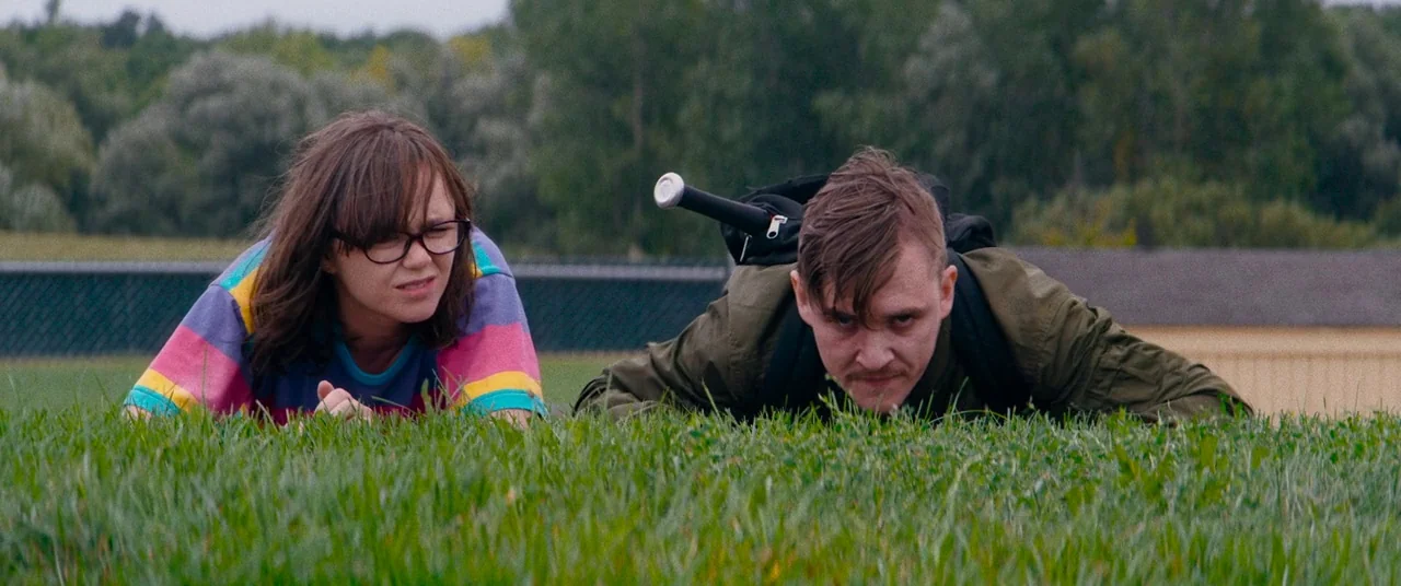 Dinner in America (2020) movie still: medium — Two individuals lie flat in the grass of an open outdoor field. The man has a baseball bat strapped…