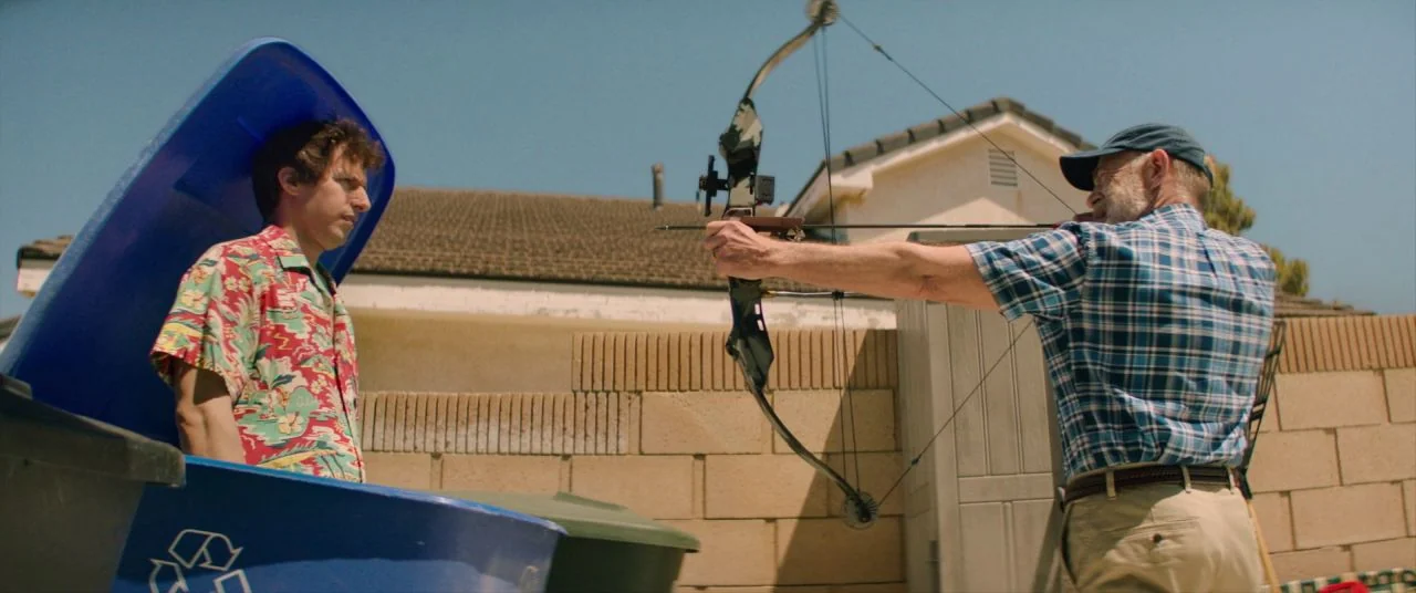 Palm Springs (2020) movie still: medium — A suburban backyard features a tan stucco wall and beige residential siding. An older man aims a…