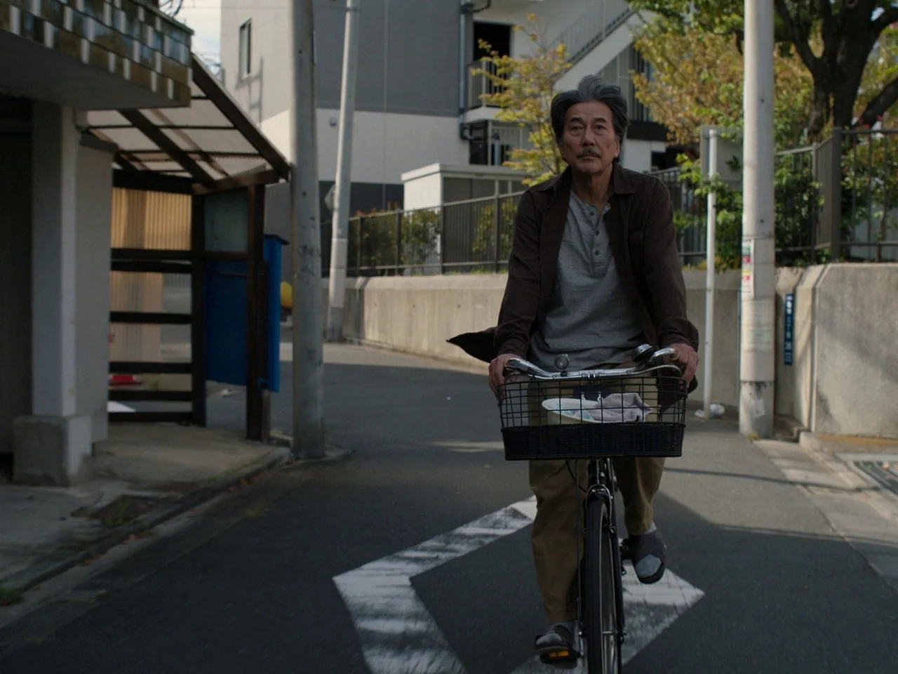 Perfect Days (2023) movie still: medium — A paved residential street features low-rise multi-story buildings and metal fences in daylight. An older…
