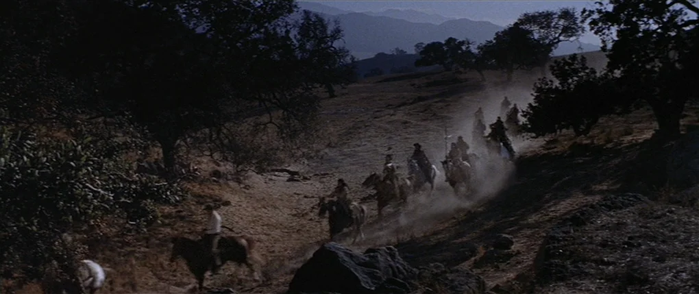 Flaming Star (1960) movie still: wide — A dry, dusty trail winds through a sparse landscape of oak trees and hills. A group of riders on horseback…