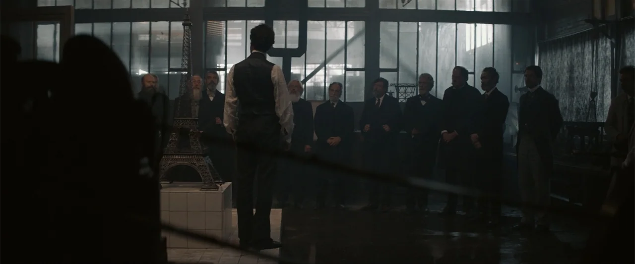 Eiffel (2021) movie still: medium — A dimly lit 19th-century workshop features large gridded industrial windows and high ceilings. A man stands with…