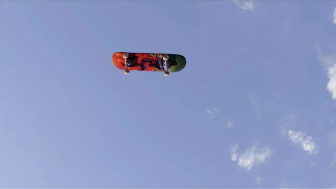 Jinxed (2013) movie still: wide — A clear blue sky with light clouds serves as the background for the shot. A colorful skateboard is suspended in…
