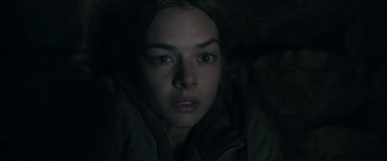 Azrael (2024) movie still: closeup — A dark space with indistinct stone-like textures visible in the shadowed background. A young woman's face is…