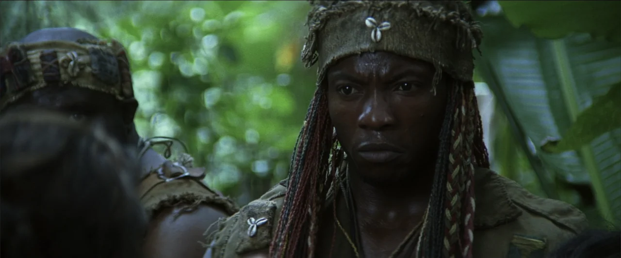 Blood Diamond (2006) movie still: medium — A dense green jungle setting with lush foliage and dappled sunlight filtering through. Two men wearing…