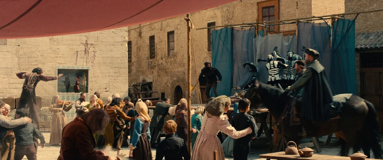 Benedetta (2021) movie still: wide — A bustling medieval square features stone buildings with a makeshift stage and blue drapes. Performers in…