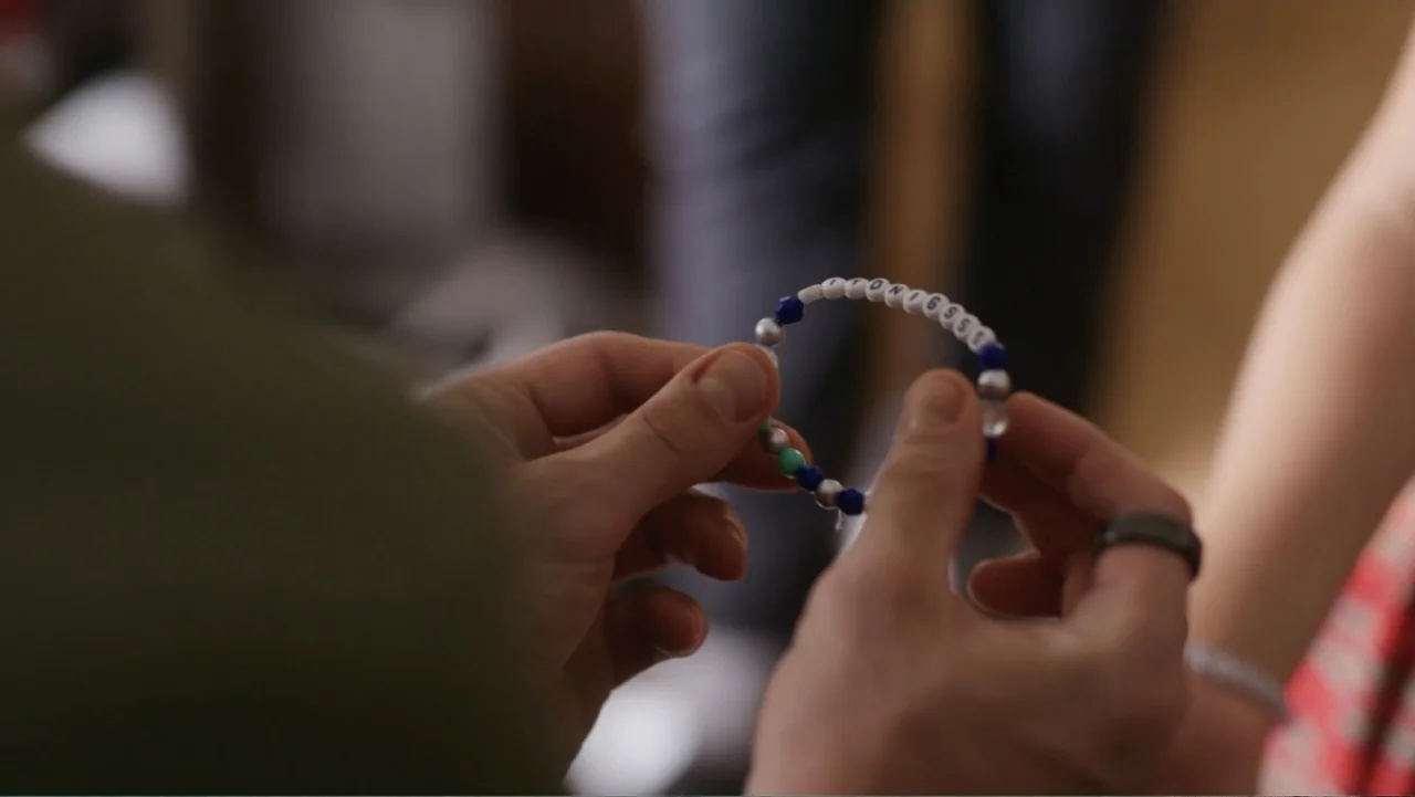 Christmas in the Spotlight (2024) movie still: closeup — A close-up view shows two sets of hands holding a small, colorful beaded bracelet. The…