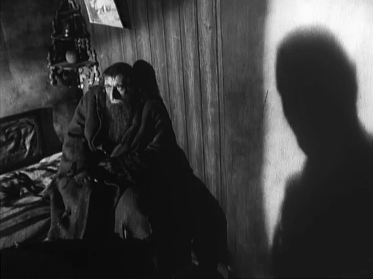 Oliver Twist (1948) movie still: medium — A dimly lit room with wood-paneled walls features a bed and a wall shadow. The bearded man sits on the…