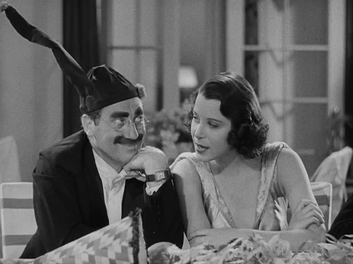 A Night at the Opera (1935) movie still: medium — A well-lit interior space with visible dining chairs and party decor on the table. A man with a…