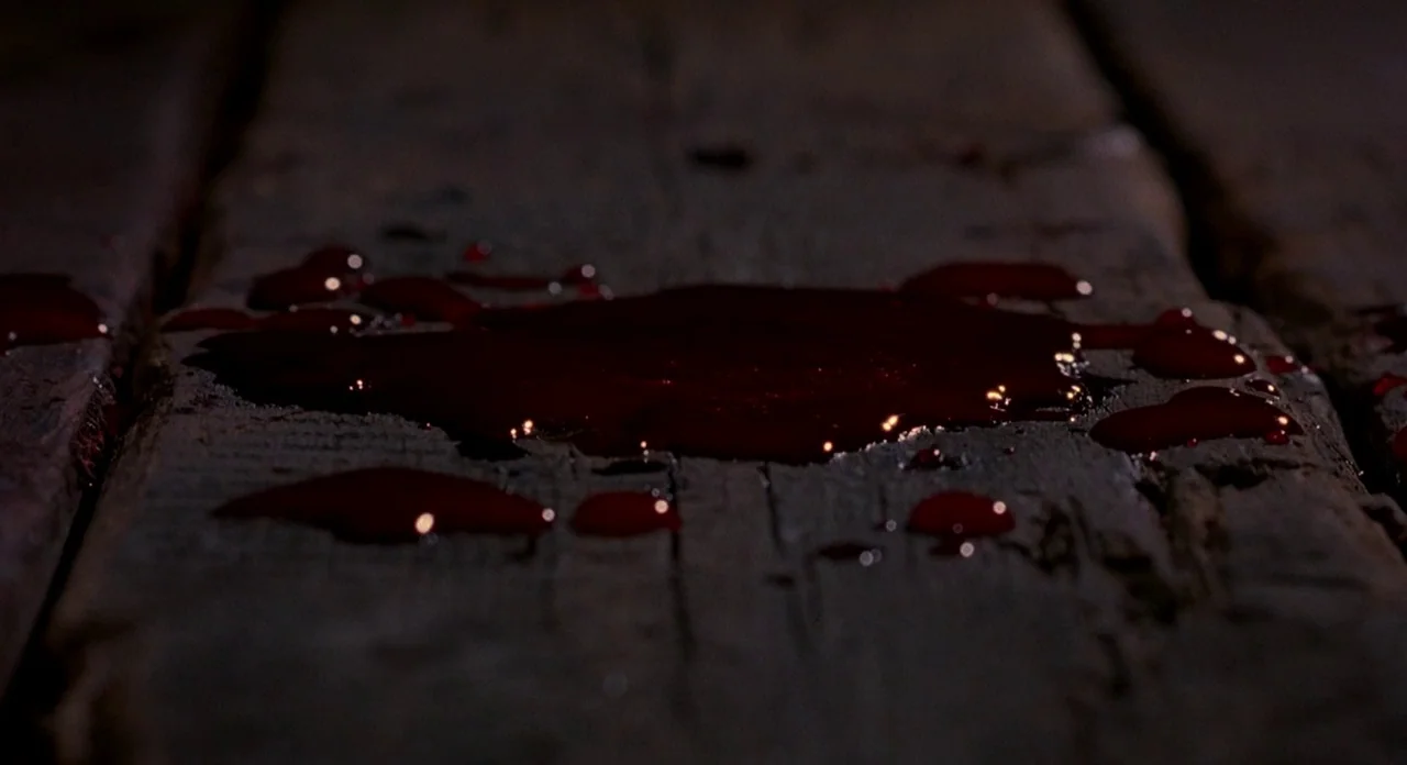 Hellraiser (1987) movie still: closeup — Dark, weathered wooden floor planks are visible in low lighting. A dark red liquid pools on the wood,…