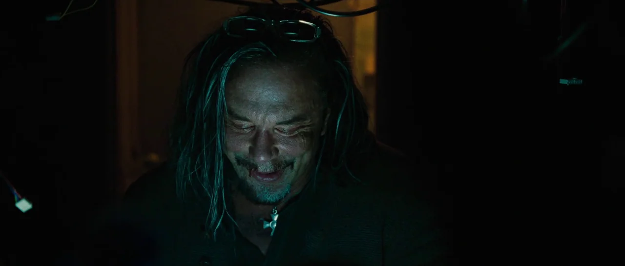 Iron Man 2 (2010) movie still: closeup — A dimly lit workspace surrounds a man with long hair illuminated by screen glow. The subject looks down…