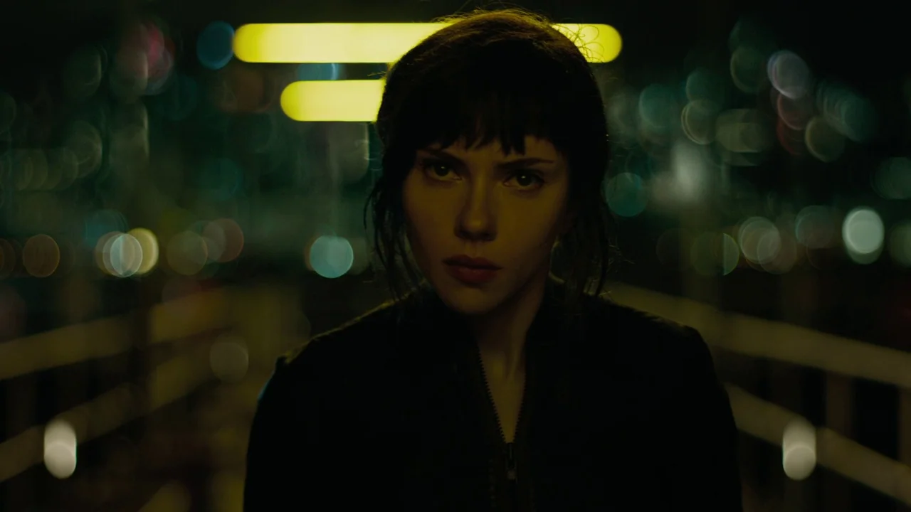 Ghost in the Shell (2017) movie still: closeup — A person stands at night on a bridge with blurry city lights behind them. The figure looks directly…