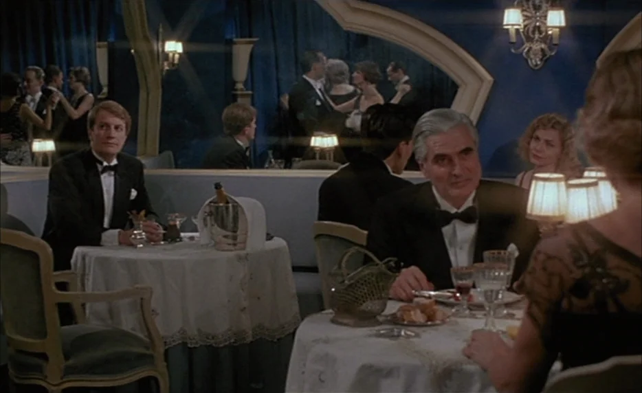 Mélo (1986) movie still: medium — An upscale dining room with blue curtains and large mirrors reflecting other patrons. A man in a tuxedo sits at a…