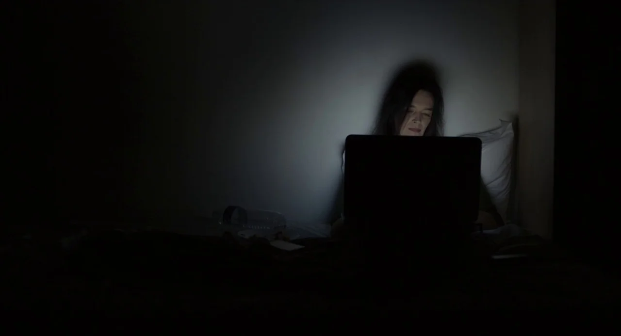 Happy End (2017) movie still: medium — A dark room features a person sitting on a bed with a glowing laptop. The screen light illuminates the…