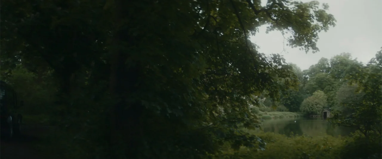 Black Bag (2025) movie still: wide — A dense grove of dark trees stands next to a small, still lake. A wooden structure is partially visible on the…