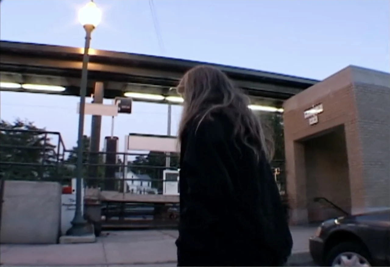 Dope Sick Love (2005) movie still: medium — A train station platform at dusk features a streetlamp and a brick structure. A woman with long hair…