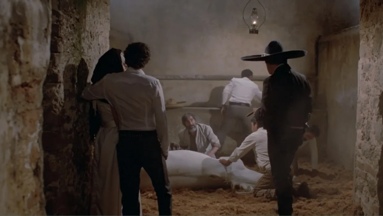 Toña Machetes (1985) movie still: fullbody — A stone-walled interior with straw on the floor and a hanging oil lamp. Several men in period attire…
