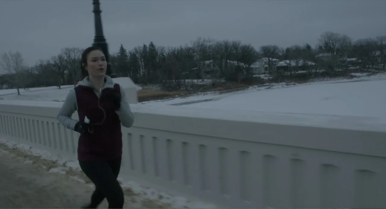 Orphan: First Kill (2022) movie still: medium — A woman jogs on a snow-covered concrete bridge during an overcast winter day. Beyond the bridge are…