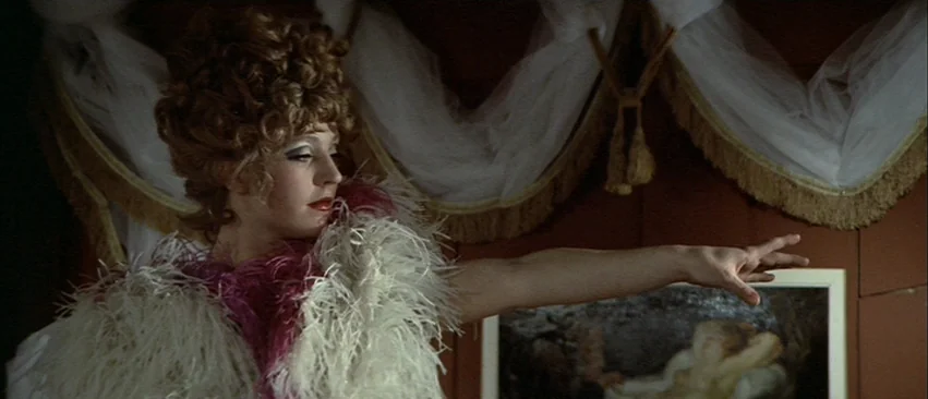 Whity (1971) movie still: medium — A dimly lit room with draped fabric curtains against a terracotta-colored wall. A woman in a feather boa extends…